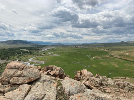 Blick auf eine weitläufige grüne Landschaft mit bewölktem Himmel und Felsen im Vordergrund.