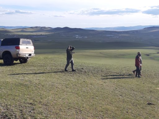 Zwei Personen und ein Auto auf einer weiten, offenen Graslandschaft.