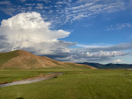 Weite Landschaft mit grünen Wiesen, einem kleinen Fluss und dramatischem Himmel.