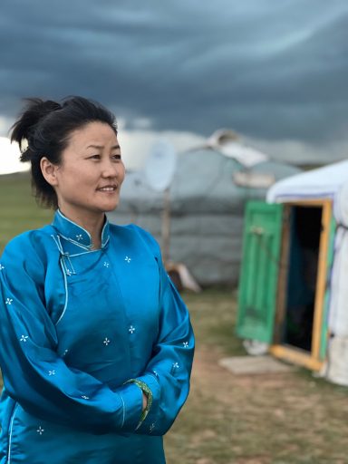 Frau in blauer traditioneller Kleidung vor einer Jurte unter bewölkten Himmel.